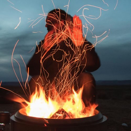 A person enjoying warmth by a vibrant campfire at twilight, sparks flying into the night sky.