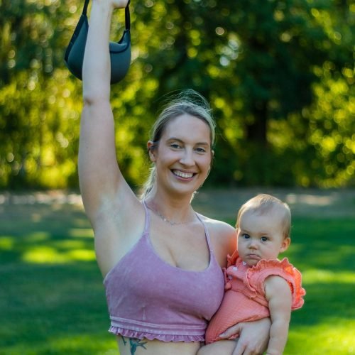 Smiling woman exercising outdoors with baby, holding a kettlebell.